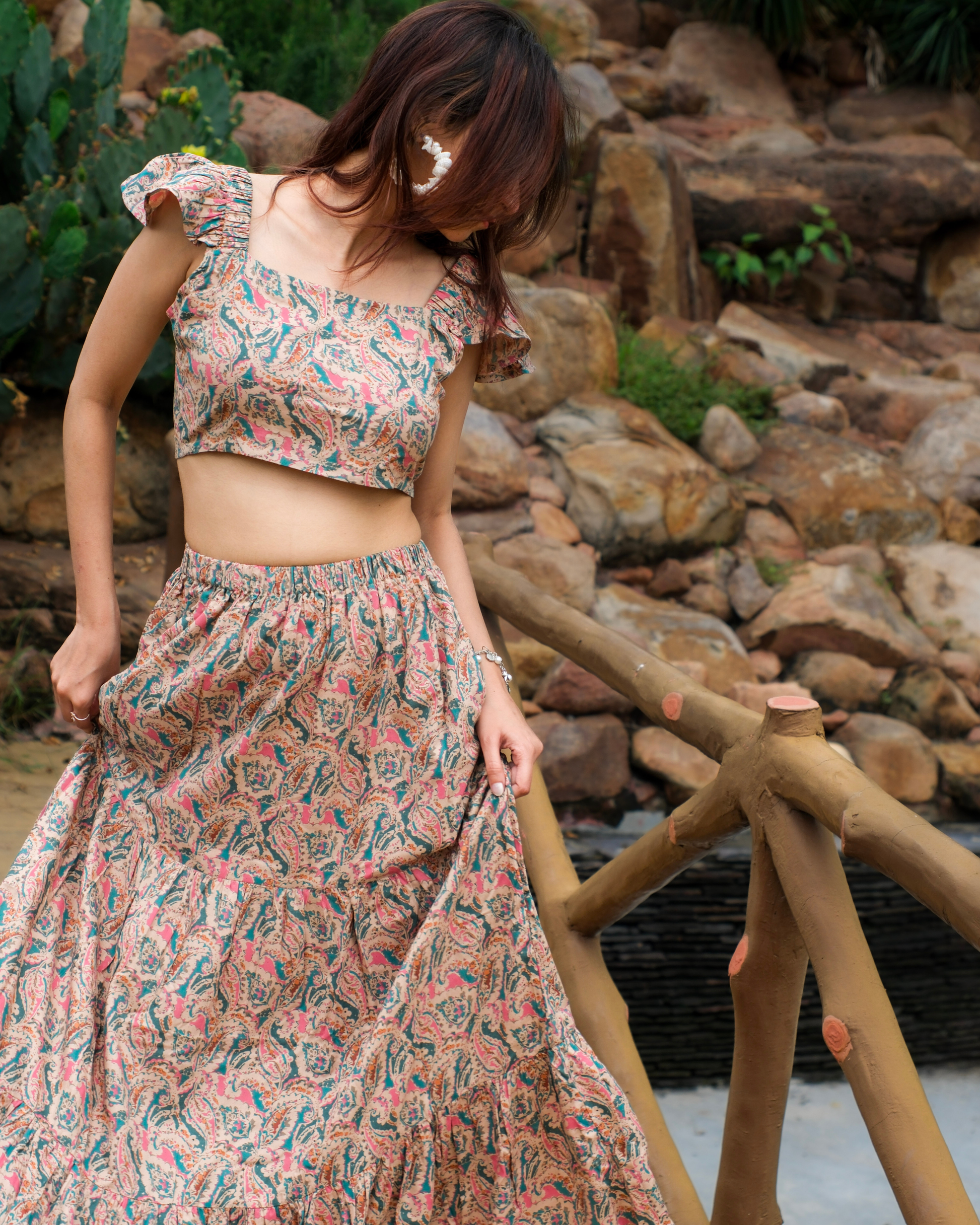women in a beige paisley printed crop top and maxi skirt set which has pockets standing on bridge. 