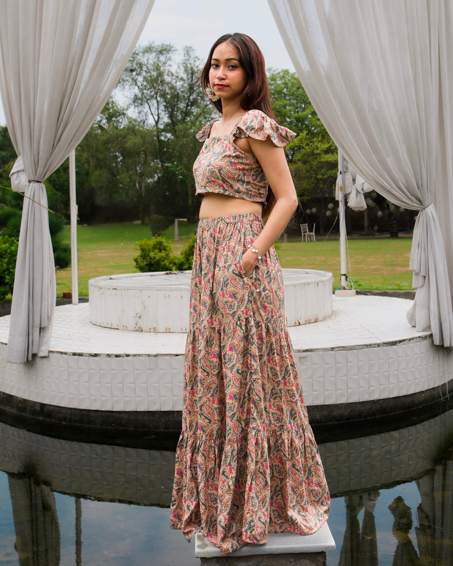 women in a beige paisley printed crop top and maxi skirt set standing near a water body decorated with curtains 