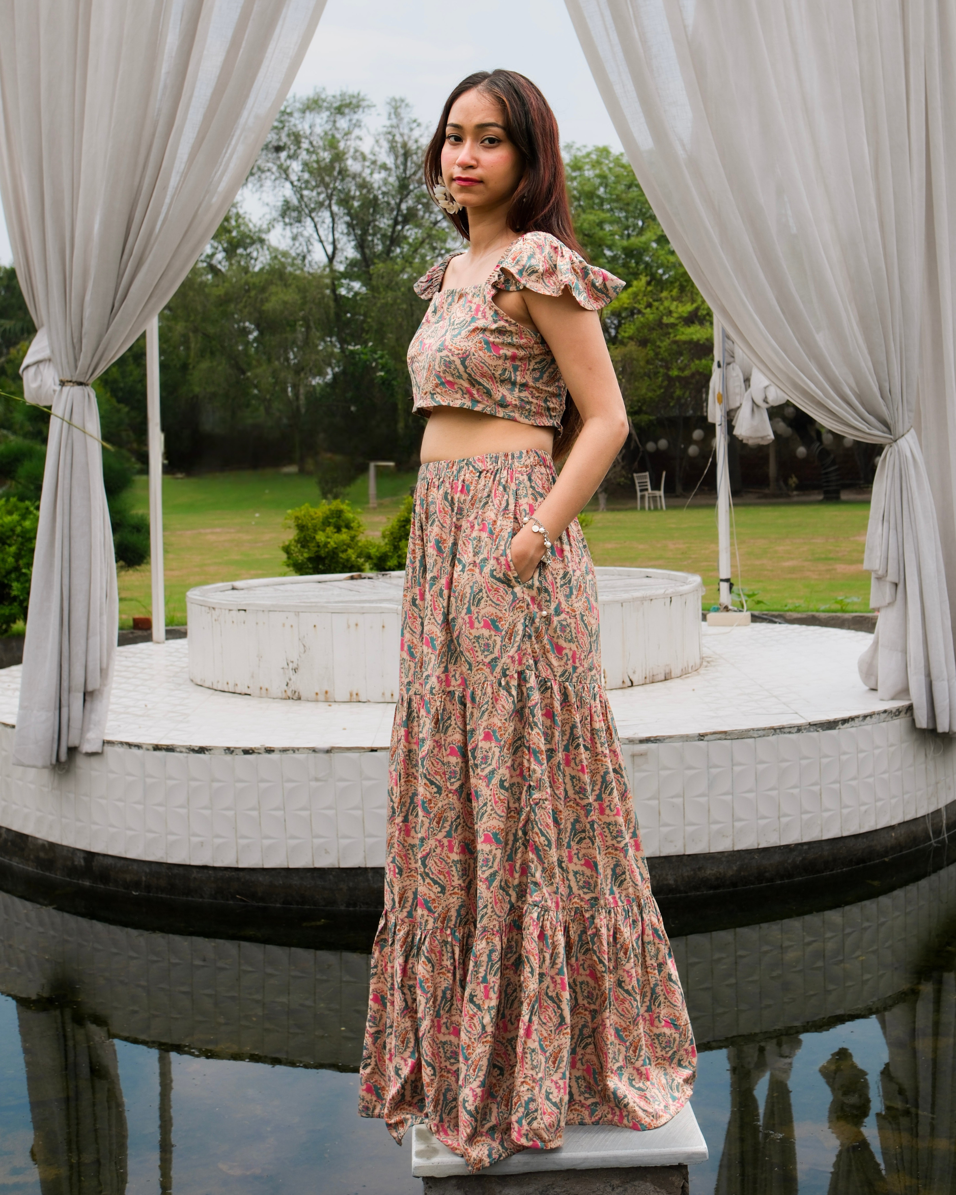 women in a beige paisley printed crop top and maxi skirt set standing near a water body decorated with curtains 