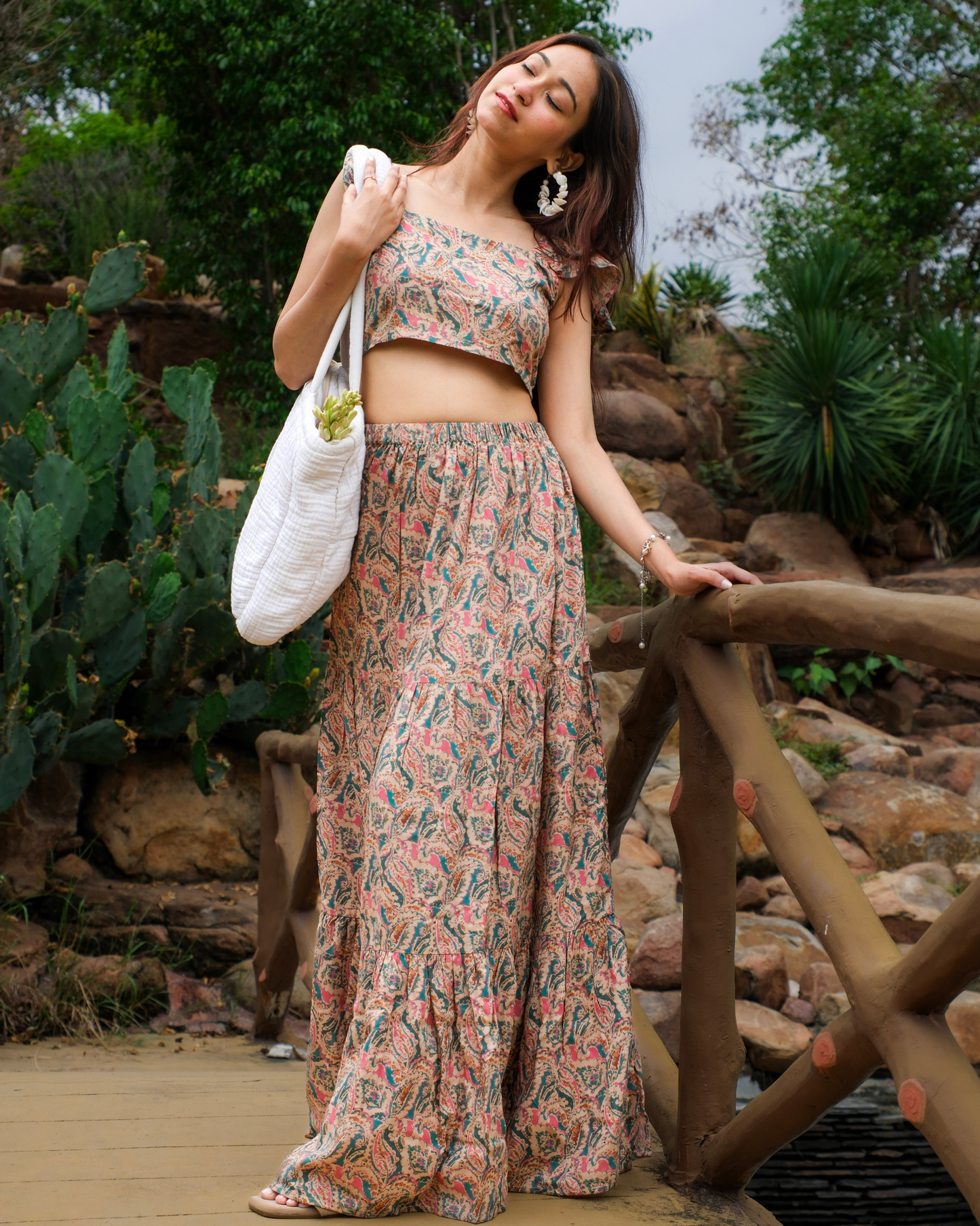 women in a beige paisley printed crop top and maxi skirt set standing on bridge with her tote bag and flowers.