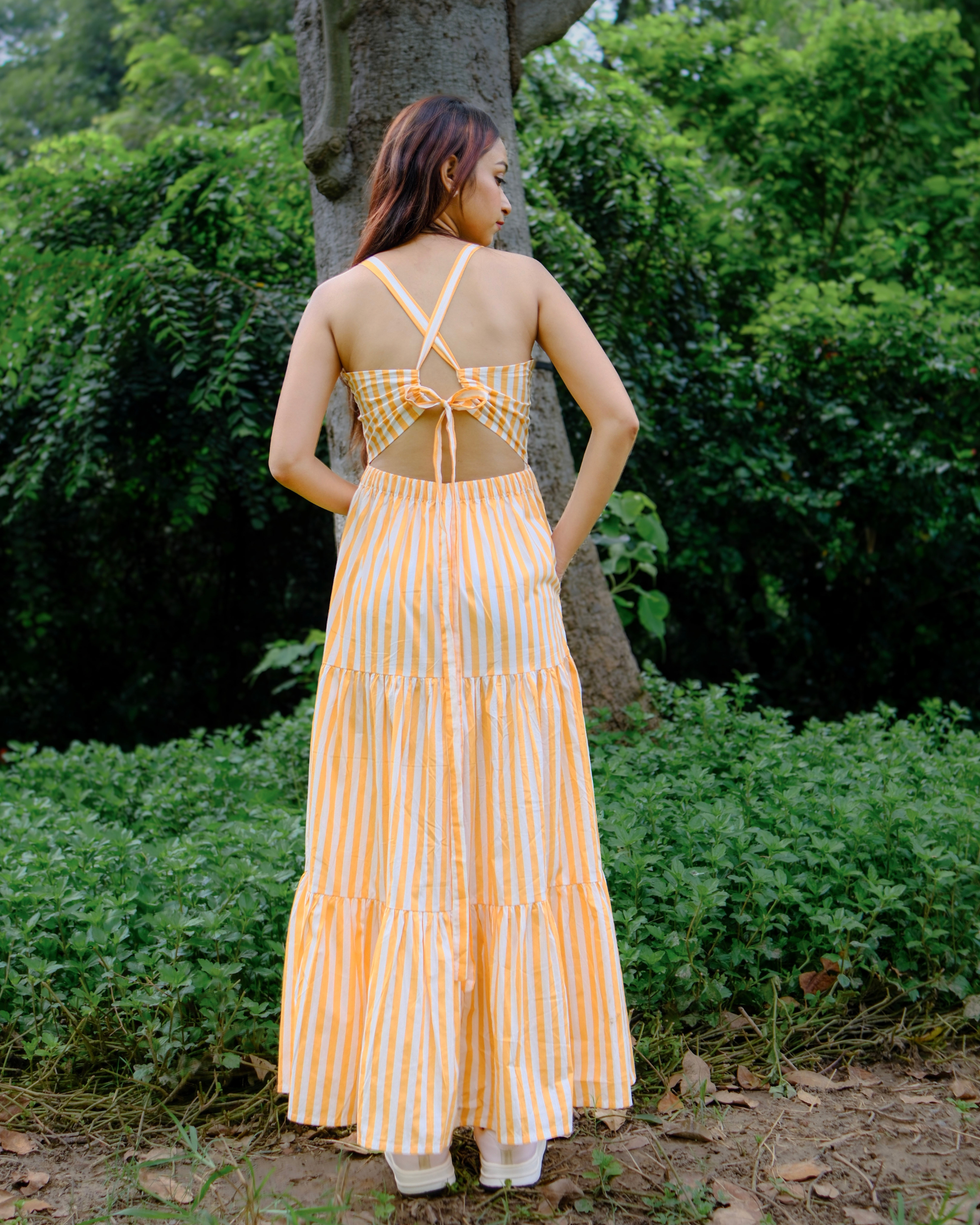 women wearing a white and orange stripe pure cotton tiered maxi dress from moodora standing in a park showing the back design of the dress which has back tie and elastic at waist.