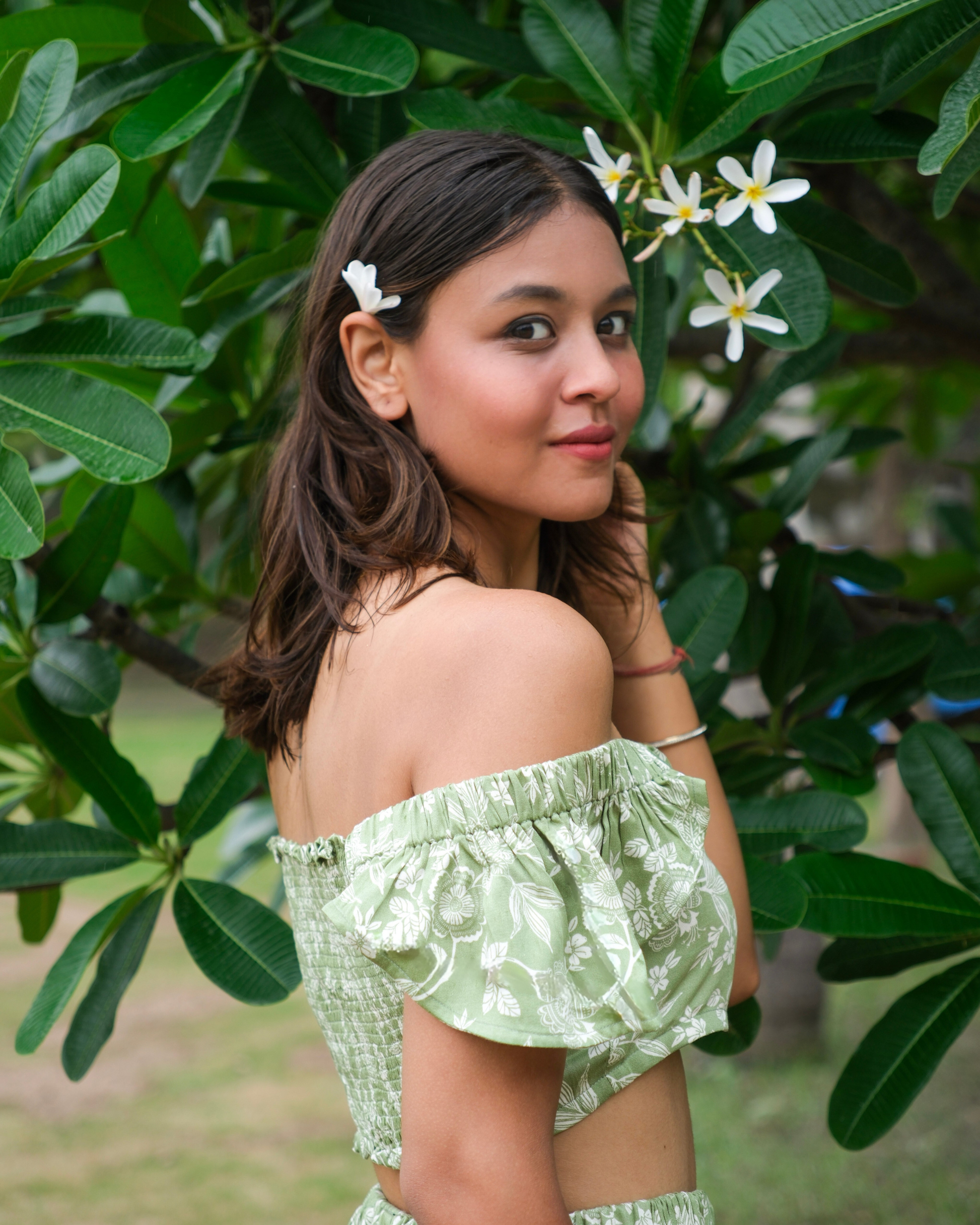 Woman in a green crop top and skirt standing in front of green foliage with white flowers.