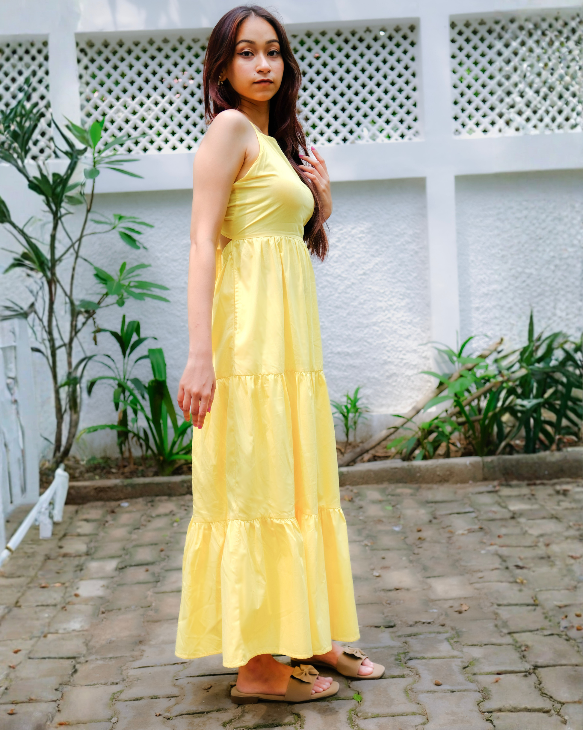 Woman in a mango yellow maxi dress by moodora standing outdoors with plants and a white wall in the background.
